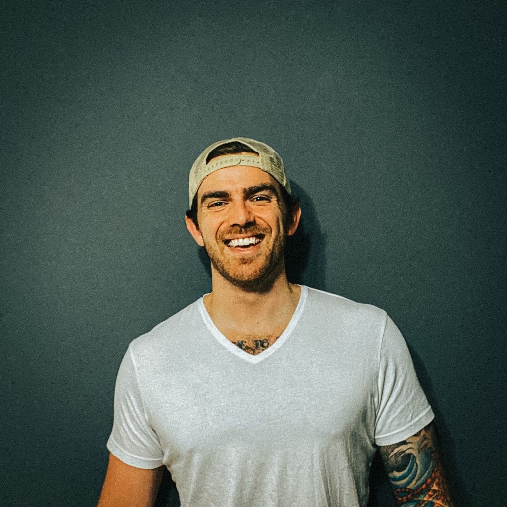 A man in a white t-shirt with tattoos and a hat creating web designs and developing websites.